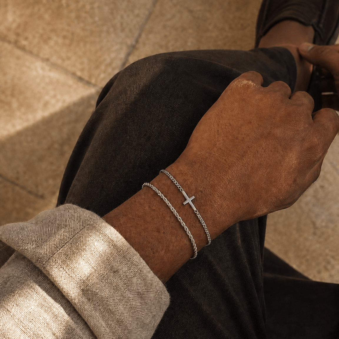 A close-up lifestyle shot of a person's wrist wearing two delicate silver chain bracelets, one of which features a small cross charm. The person is wearing dark trousers, a light beige knit sweater, and a dark brown loafer, seated on a tiled floor.